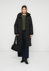 Sort pufferjakke med hætte, der har et quiltet design. Båret over en grøn sweater, parret med brede jeans og sorte ankelstøvler.