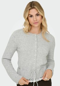 Donna con capelli biondi che indossa un cardigan leggero grigio chiaro con bottoni in perla e un papillon, sopra un top bianco con bordo in pizzo.