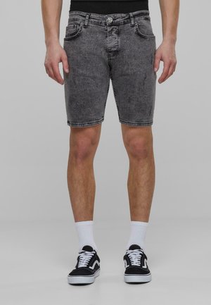 Zwarte denim short met een licht beschadigde textuur, vijf zakken en een knoopsluiting, gecombineerd met zwarte sneakers en witte sokken.