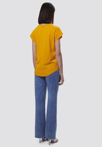 T-shirt giallo a maniche corte con orlo curvo, abbinato a jeans in denim blu a gamba larga e sandali con tacco. Vista da dietro.