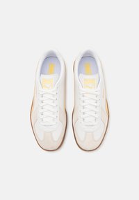 Scarpe sportive bianche in materiali scamosciati e pelle, con dettagli gialli, lacci piatti e suola in gomma. Logo visibile sulla linguetta.