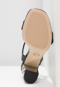 Sandale à talons hauts noirs avec un bout ouvert, tige en cuir verni, accent métallique décoratif et semelle en caoutchouc texturée.