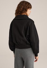 Žena s vlnitými hnědými vlasy, oblečená v černé oversized bundě a šedých kalhotách, stojící bokem proti jednoduchému béžovému pozadí.