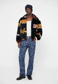 Veste en fausse fourrure noire avec des motifs orange et blancs, associée à un jean bleu à motifs. Le mannequin porte un bonnet et des chaussures noires.