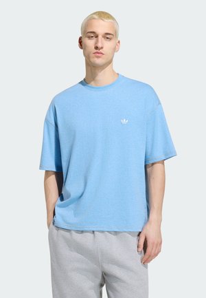 Jeune homme aux cheveux blonds courts portant un t-shirt Adidas bleu clair et un pantalon de survêtement gris clair, debout devant un fond uni.