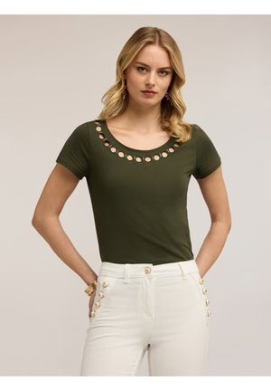 Vaaleahiuksinen nainen, jolla on oliivinvihreä t-paita pyöreillä leikkauksilla pääntiessä ja valkoiset housut, joissa on kultaiset napit.