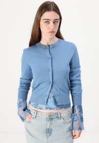 Svijetloplavi kardigan od mekog pletenja s okruglim izrezom, prednjim gumbima i dugim rukavima preko prozirne bluze s cvjetnim uzorkom.
