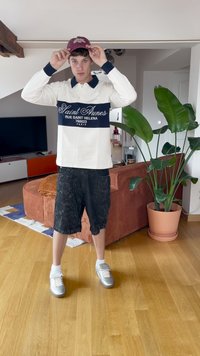 Vit långärmad polo med marinblå rand, prydd med text. Svarta mönstrade shorts och silvriga sneakers. Inomhusmiljö med grön växt.