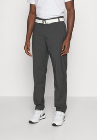 Mann som har på seg mørkegrå bukser, hvit belte, hvit T-skjorte og hvite sneakers, står på et lysegrått gulv mot en hvit vegg.
