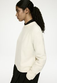 Cremefarbener, gestrickter Pullover mit rundem Ausschnitt, gerippten Bündchen und cropped Passform. Die Textur wirkt weich und leicht dick.