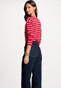 Langärmliges Oberteil mit roten und weißen Streifen und gerafften Details an den Schultern, kombiniert mit hochgeschnittenen dunkelblauen Jeans und mehreren silbernen Armbändern.