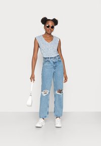 Top azul sem mangas com padrões florais, jeans rasgados de cintura alta em azul claro, tênis brancos e uma pequena bolsa branca.