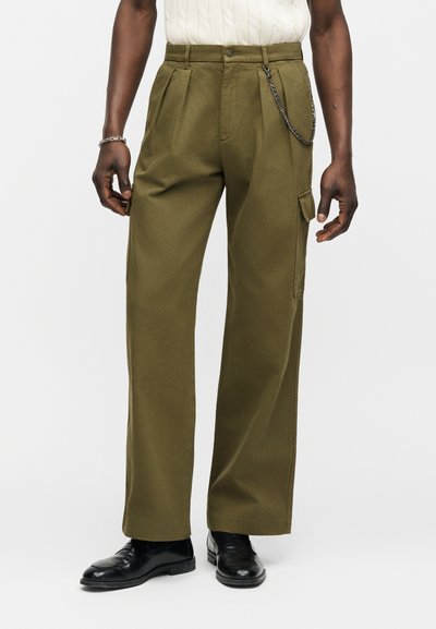 Άνδρας που φοράει χακί παντελόνι καμπάνα με πιέτες, αλυσίδα να κρέμεται από το θηλάκι της ζώνης, μαύρα παπούτσια και λευκό πλεκτό τοπ.