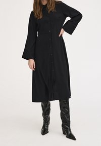 Abito midi nero a coste con chiusura a bottoni, maniche a campana lunghe e vita definita. Indossato con stivali a tacco alto neri lucidi con effetto coccodrillo.