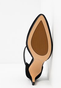 Chaussure à talon haut en daim noir à bout pointu vue par dessous avec une semelle beige et une semelle antidérapante marron texturée au centre.