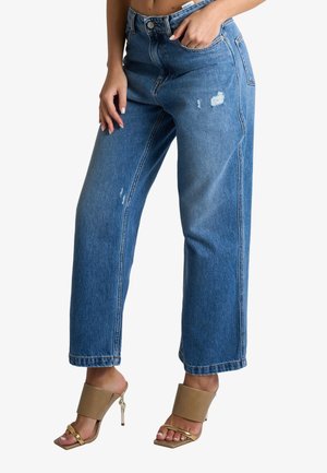 Donna che indossa jeans blu a vita alta, gamba dritta con leggero effetto consumato, abbinati a sandali beige con tacco alto su sfondo bianco.