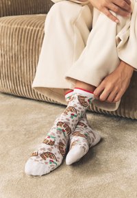 Calzini bianchi con stampa leopardata e motivo di agrifoglio, con polsino a righe rosse, indossati ai piedi di una persona, abbinati a pantaloni color crema.