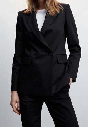 Mujer con blazer negro de doble botonadura hecho a medida sobre una camiseta blanca y pantalones negros a juego, de pie con una mano en el bolsillo.
