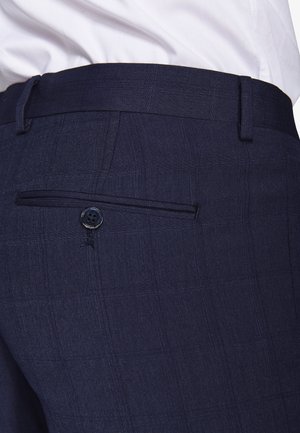 Nærbillede af marineblå bukser med en knappet baglomme, båret med en hvid skjorte, der er stoppet ind.