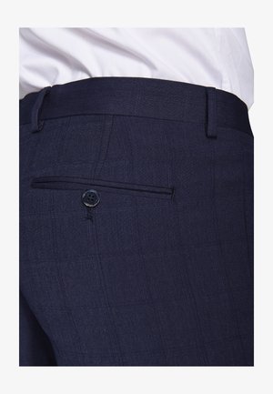Primo piano di pantaloni da abito blu navy con tasca posteriore con bottone, indossati con una camicia bianca infilata dentro.