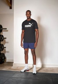 Sort t-shirt med hvid "PUMA" logo, blå sportsshorts, hvide sneakers med røde og sorte detaljer, stående i et fitnessrum med vægte.