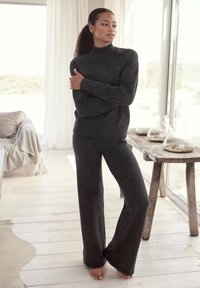 Gemütlicher, dunkelgrauer Strickpullover mit Rollkragen, kombiniert mit weit geschnittenen Hosen aus weichem, strukturiertem Stoff. Das Model steht drinnen neben einem Holztisch.