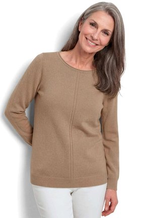 Femme souriante aux longs cheveux gris, vêtue d'un pull beige à manches longues et d'un pantalon blanc, debout sur un fond blanc.