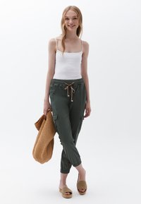 Olijfgroene cargobroek met een elastische tailleband en trekkoord, gecombineerd met een witte tanktop. Het model houdt een tan geknoopte tas vast en draagt geweven sandalen.