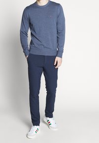 Maglione a maniche lunghe blu navy con scollo rotondo, abbinato a pantaloni slim-fit blu scuro. Sneakers bianche con accenti verdi e rossi.
