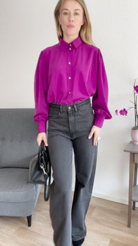 Magenta bluse med krage og frontknapper, kombinert med høyt liv svarte jeans. Holder en svart håndveske, står ved en grå lenestol.