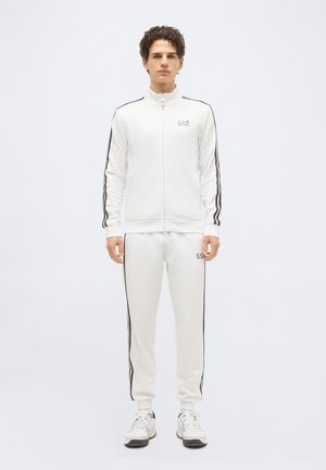 Νεαρός άνδρας που στέκεται, φορώντας λευκή φόρμα EA7 με λεπτομέρειες σε μαύρο και κόκκινο ρίγα και λευκά αθλητικά παπούτσια, σε απλό φωτεινό φόντο.