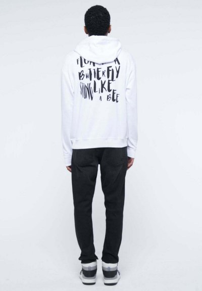 Felpa bianca con una frase stampata in nero sulla schiena, abbinata a pantaloni scuri e sneakers bianche, evidenziando un look casual e rilassato.