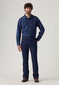 Camisa de denim azul oscuro con mangas largas y bolsillos en el pecho, combinada con jeans ajustados de denim azul oscuro y botas de vaquero marrones.