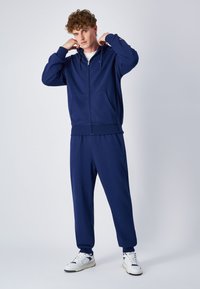 Navyblauer Reißverschluss-Hoodie mit Fronttaschen, kombiniert mit navyblauen Jogginghosen. Der Stoff hat eine glatte Textur. Weiße Sneaker mit schwarzen Akzenten.