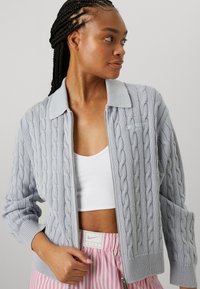 Light gray, cable-knit zip-up cardigan with a ribbed collar and cuffs. Features subtle branding on the chest and relaxed fit.