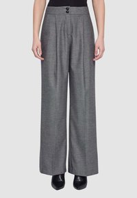 Donna che indossa pantaloni grigi a vita alta e gamba larga con pieghe frontali e stivaletti neri su uno sfondo semplice.