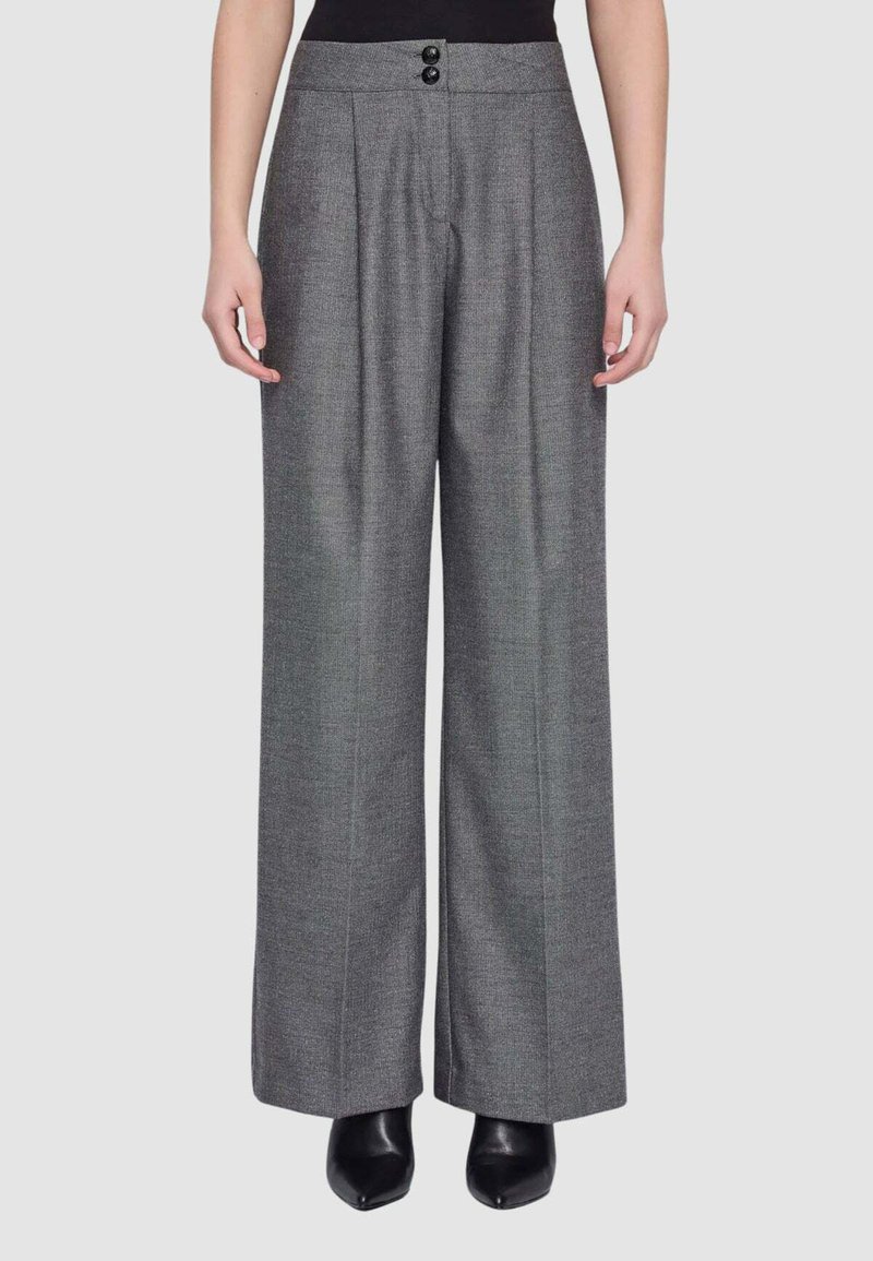 Donna che indossa pantaloni grigi a vita alta e gamba larga con pieghe frontali e stivaletti neri su uno sfondo semplice.