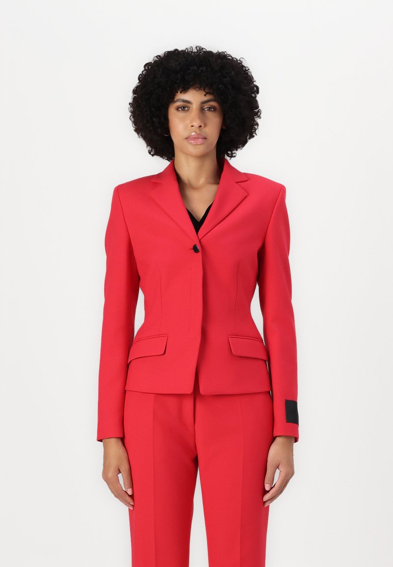 Veste rouge et pantalon assorti, fabriqués en tissu texturé. Coupe ajustée, avec une fermeture à un bouton et des détails de poches.