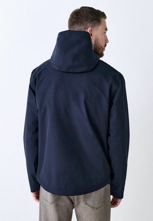 Mørkeblå vandtæt jakke med hætte, blød tekstur, front sømdetalje og afslappet pasform, båret over lysfarvede jeans.
