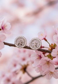 Orecchini rotondi in argento con un design centrale a forma di albero, circondato da piccole gemme trasparenti, su uno sfondo di fiori di ciliegio.