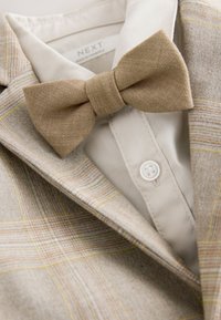 Light tan plaid suit jacket with subtle yellow accents, paired with a beige bow tie and a cream-colored dress shirt buttoned at the collar.