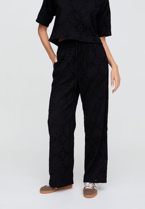 Frau trägt schwarze Spitzen-Hosen mit weitem Bein, kombiniert mit einem passenden kurzärmligen Oberteil und braunen Schnürschuhen vor neutralem Hintergrund.