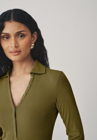 Olivengrøn ribstrikket langærmet top med krave og knaplukning. Glat tekstur med tætsiddende pasform.