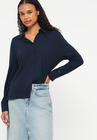 Camisa azul-marinho de botão, com um corte descontraído, mangas longas e gola, combinada com uma saia de ganga azul-clara.