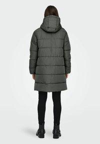 Person iført en lang, mørkegrøn, quiltet hættejakke, sorte bukser og sorte ankelstøvler, der står med ryggen til mod en hvid baggrund.
