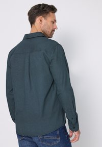 Tmavě zelená košile s dlouhým rukávem s jemným texturovaným vzorem, manžetami na knoflíky a záhybem na zádech. Kombinováno s modrými džínami.