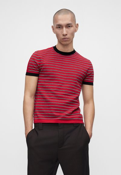 Jeune homme aux cheveux courts portant un t-shirt à manches courtes rayé rouge et noir et un pantalon noir, les mains dans les poches, sur un fond uni.