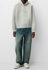 Ljust grå huvtröja med känguruficka och präglad text, i kombination med löst sittande blå jeans och vita sneakers.