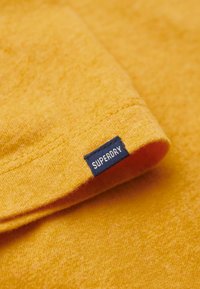 Gul strikstof med en blød tekstur, der har en foldet ærme og et blåt mærke med teksten "SUPERDRY" syet på kanten.