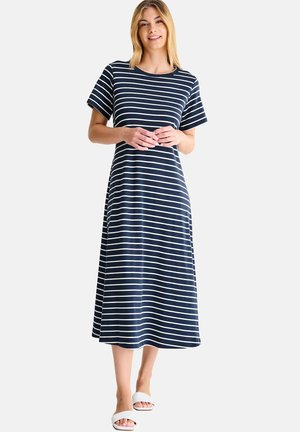 Femme portant une robe midi à rayures horizontales bleu marine et blanches, à manches courtes, avec des sandales blanches ouvertes aux orteils, debout et souriante.